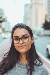 A confident woman wearing glasses smiles subtly while standing on an urban street with blurred background.