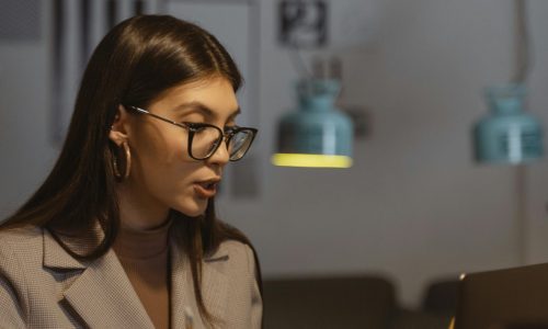 A young businesswoman working late at her desk, engaged in an online meeting with a laptop, notebook, and smartphone.