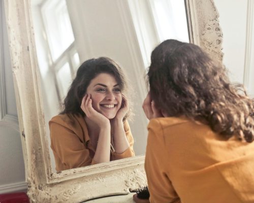 A cheerful woman smiles at her reflection in a vintage-style mirror, exuding positivity and warmth.